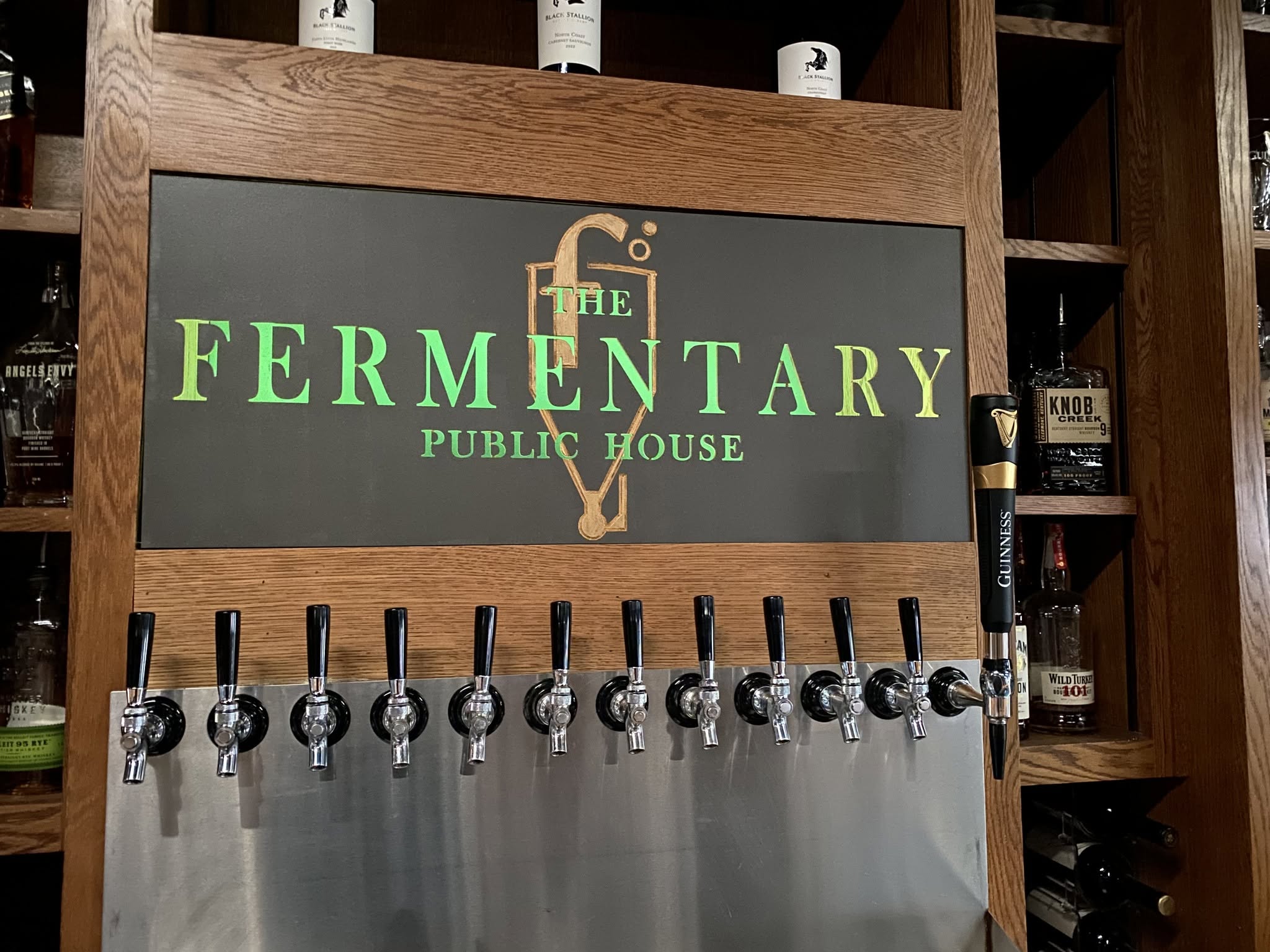 The carved Fermentary Public House sign above the tap wall, lit green, with a row of taps below.